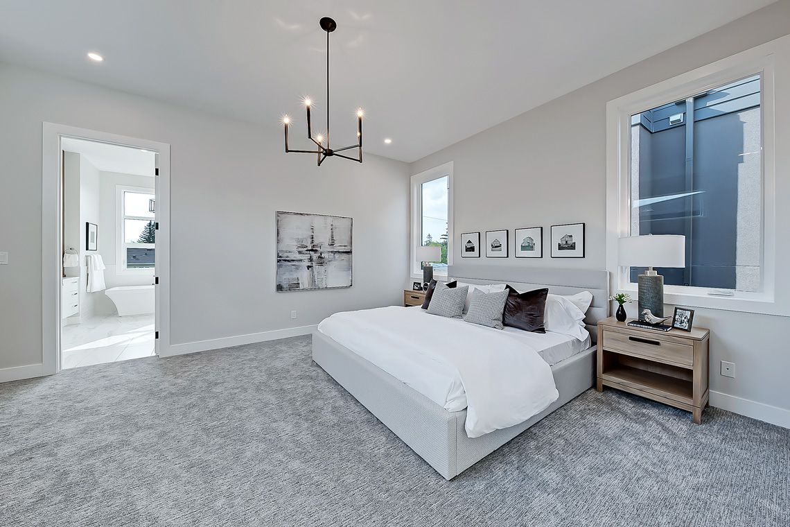 A modern bedroom with grey carpet, a white bed, a chandelier, and a large window, leading to a bathroom.