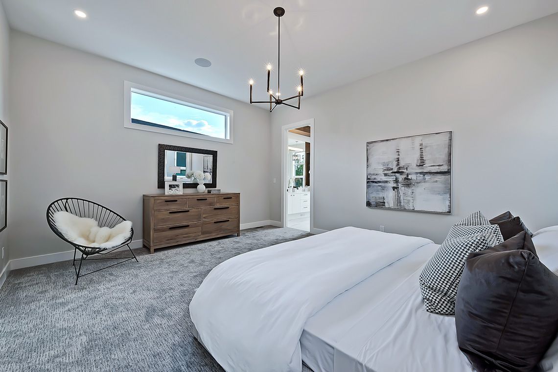 A bright, neutral-toned bedroom featuring a bed, a wooden dresser with a mirror, a chair, and a chandelier.
