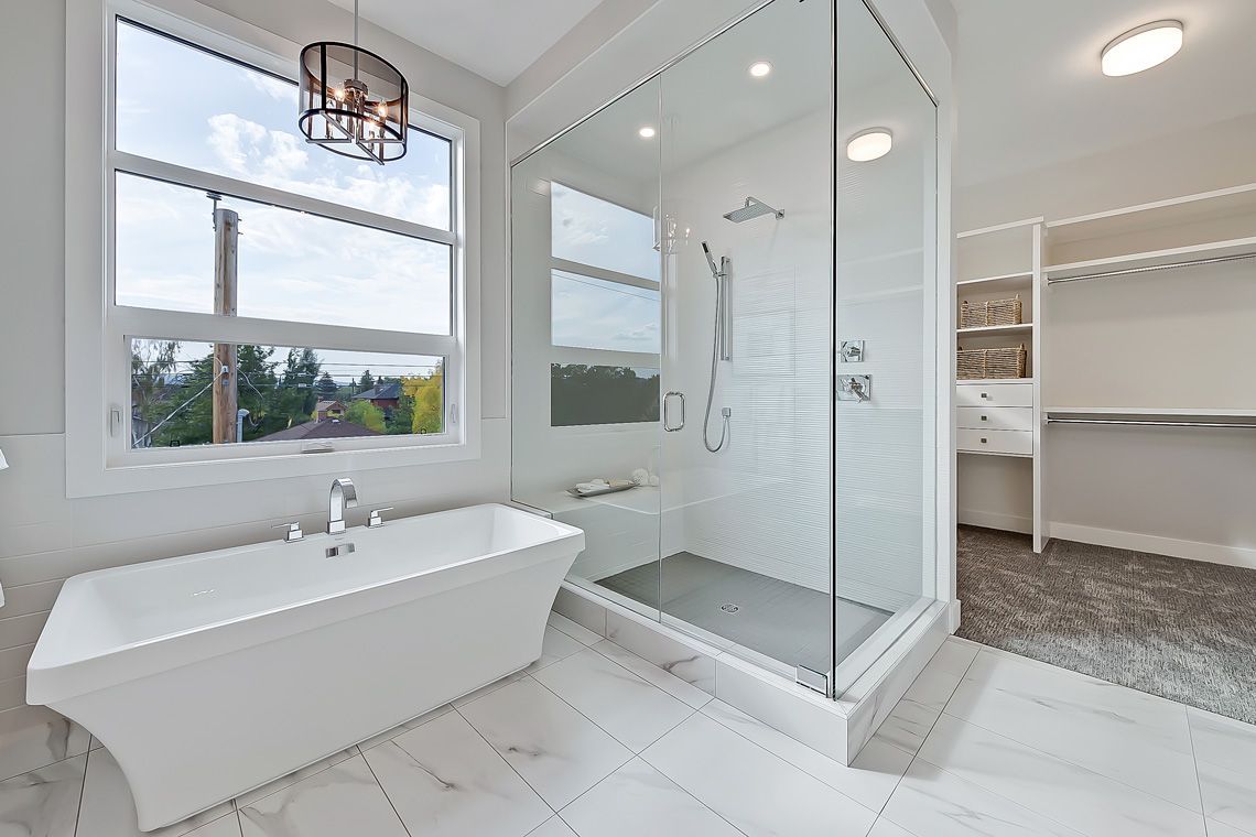 A bright, modern bathroom with a freestanding white tub, a glass-enclosed shower, and an open closet with gray carpeting.