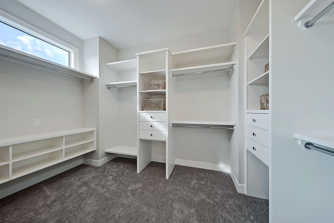 A walk-in closet featuring white wooden shelving, drawers, hanging rods, and grey carpet under a horizontal window.