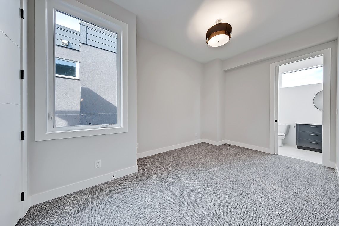 A bright, empty bedroom with grey carpet, neutral walls, a window overlooking a building, and an attached bathroom.