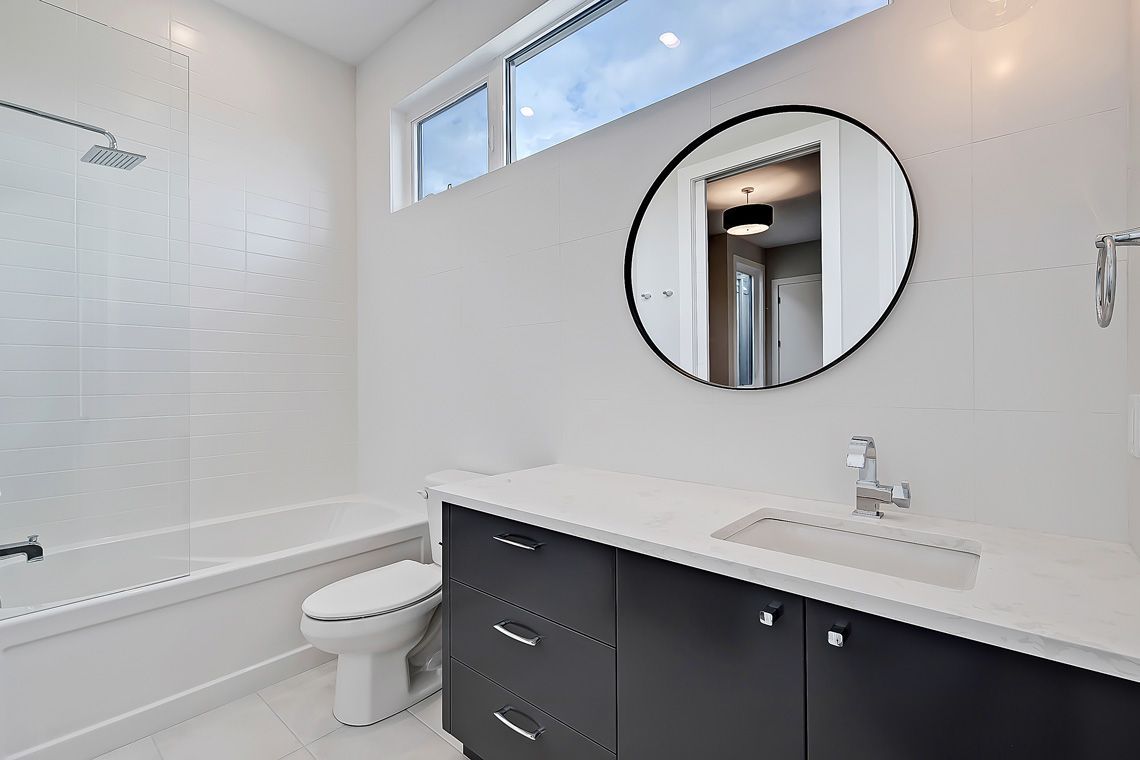 Modern bathroom with white walls, a bathtub with a glass panel, a toilet, and a dark vanity with a circular mirror.