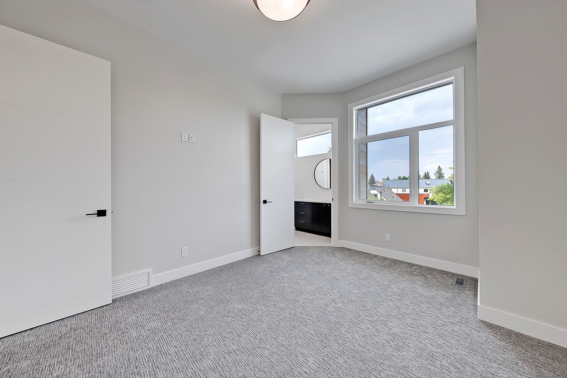 An empty, modern bedroom with grey carpet, light grey walls, a large window, and two white doors.
