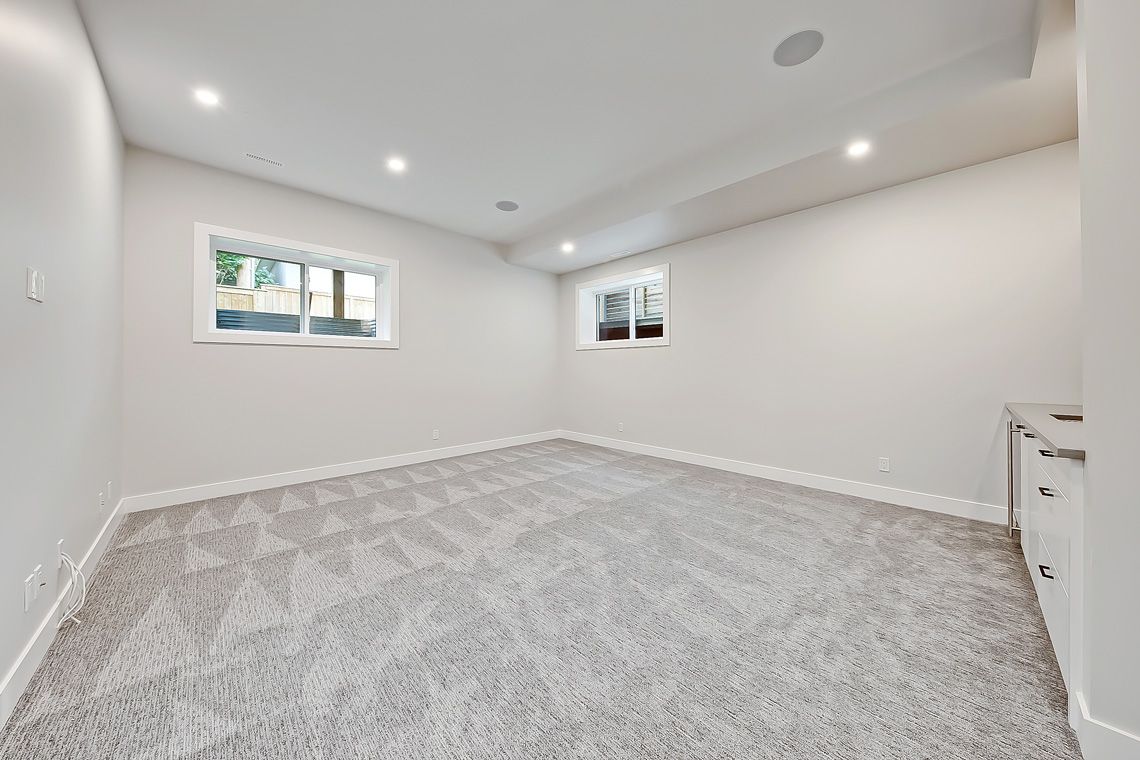 A bright, empty room with light gray carpet, white walls, recessed ceiling lights, two windows, and built-in white cabinets.
