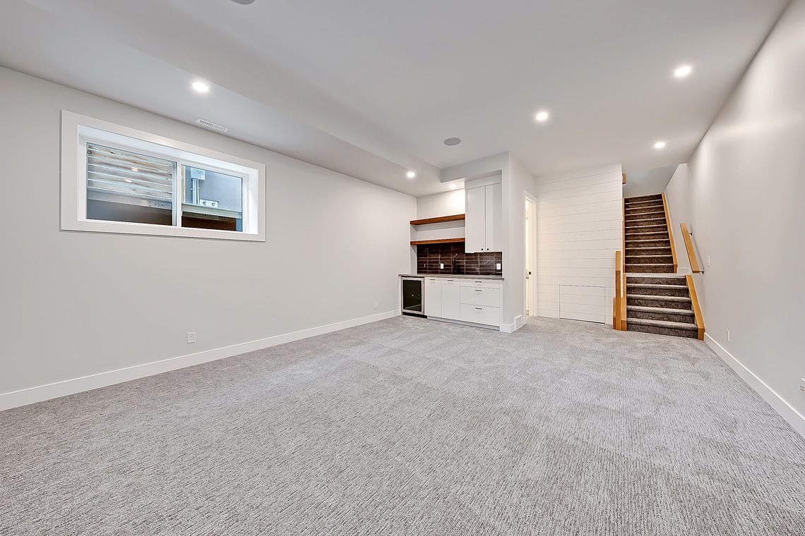 A carpeted basement room with light grey walls, a small wet bar, a window, and stairs leading to an upper level.
