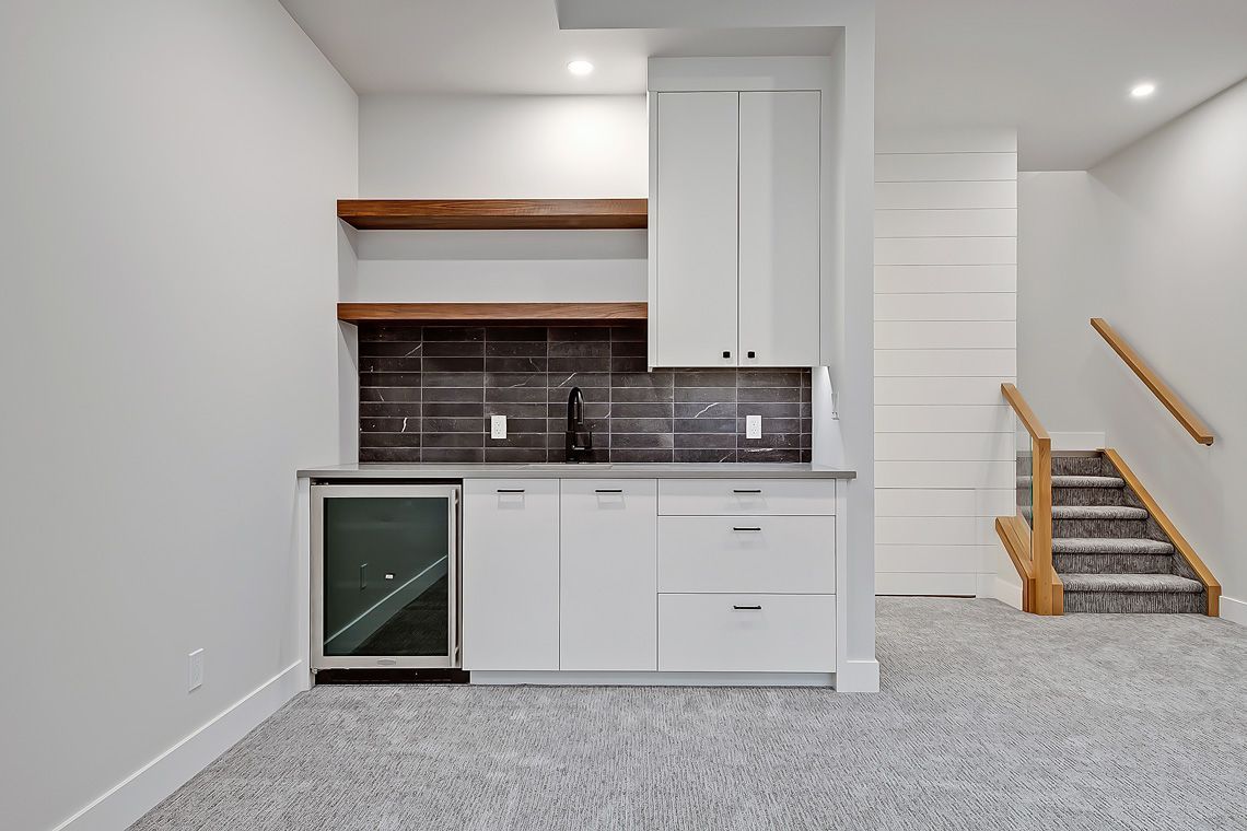 A home bar with white cabinets, wood shelves, and a dark tile backsplash, located next to a carpeted staircase.
