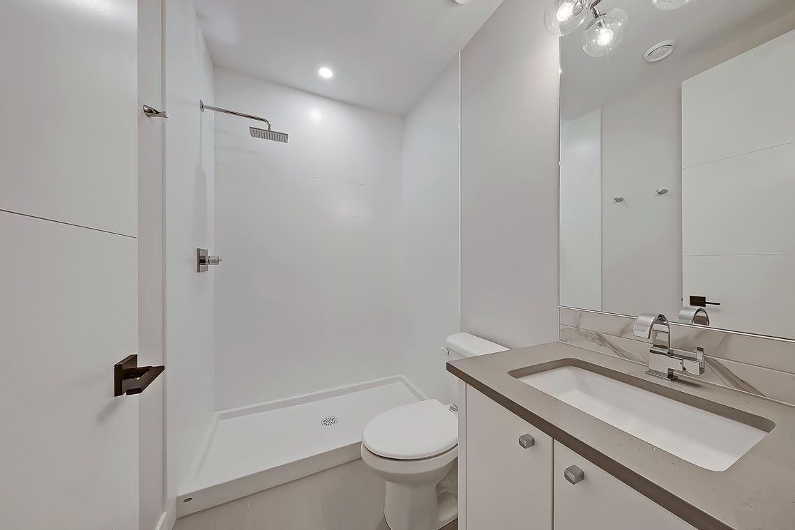 A modern bathroom with white walls, a shower stall, a toilet, and a vanity with a white sink and gray countertop.
