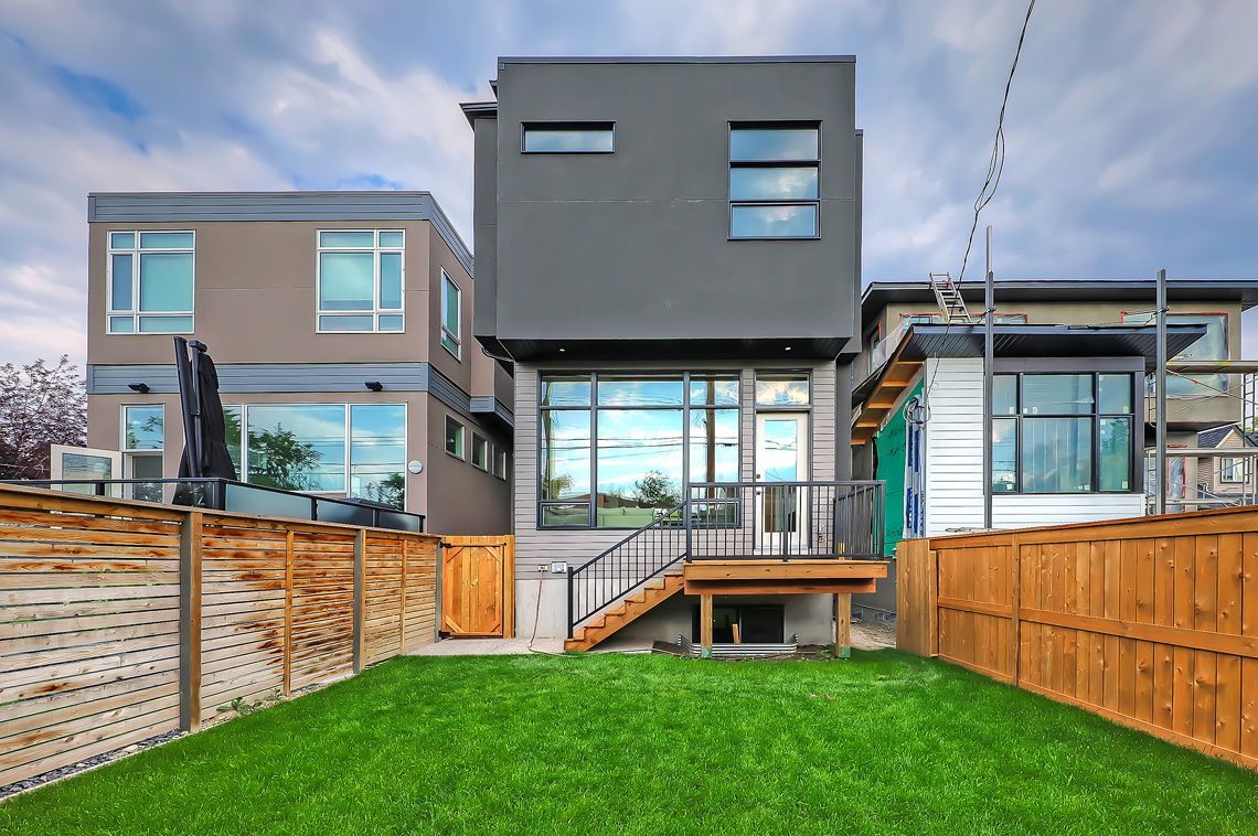 A modern house with a gray exterior, a large glass window, and a wooden deck overlooking a manicured lawn and fences.