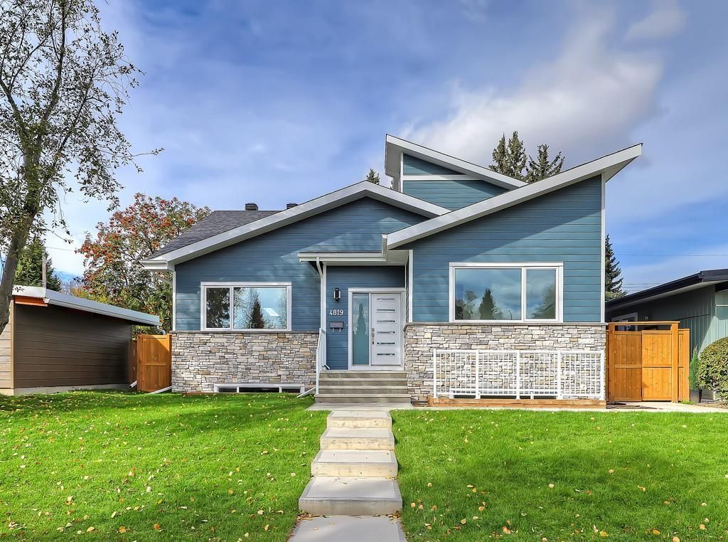 A modern blue house with a stone foundation, a multi-tiered roofline, and a central entryway under a sunny sky.