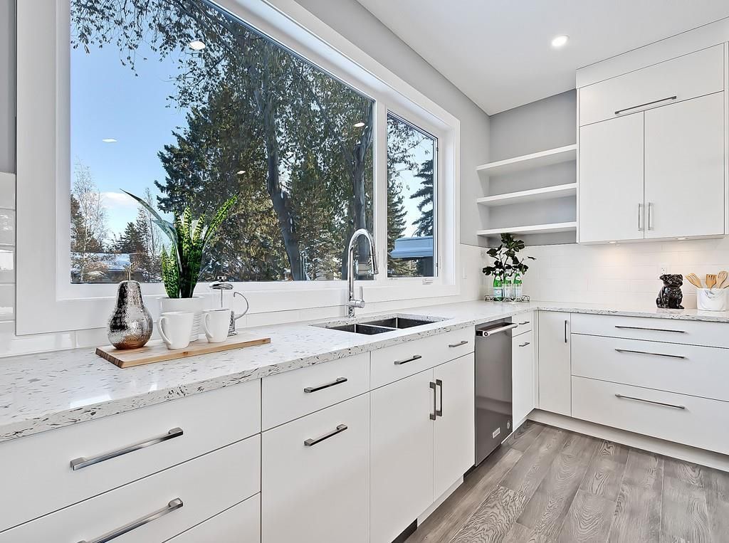 A bright, modern kitchen features white cabinets, speckled countertops, a large window, and light grey wood-look flooring.