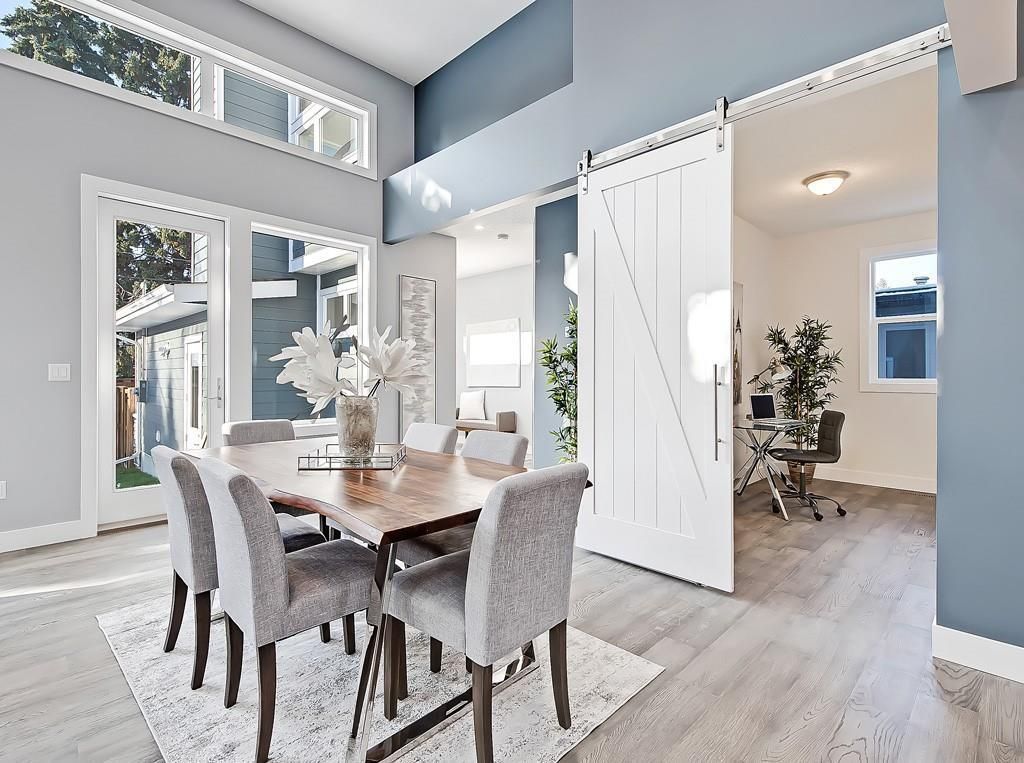 A modern dining area with a wooden table and gray chairs, opening into a home office through a white barn-style door.