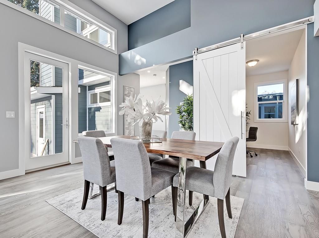 A dining room with a wood table, four gray chairs, white barn door, and large windows set against blue and white walls.