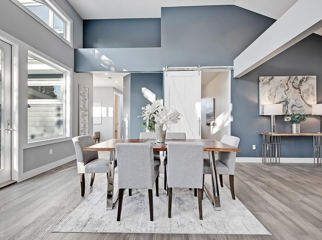 A modern dining room with a wooden table, six grey upholstered chairs, and a white barn door against a blue accent wall.