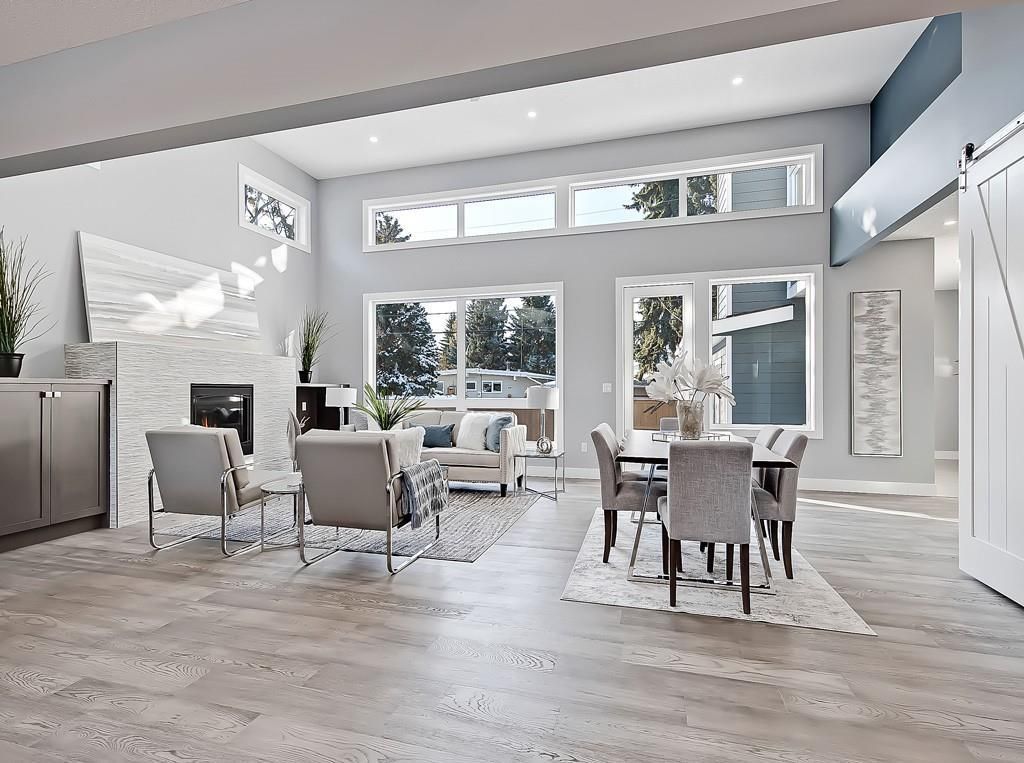 A bright, open-concept living and dining room with modern grey decor, large windows, a fireplace, and light wood floors.