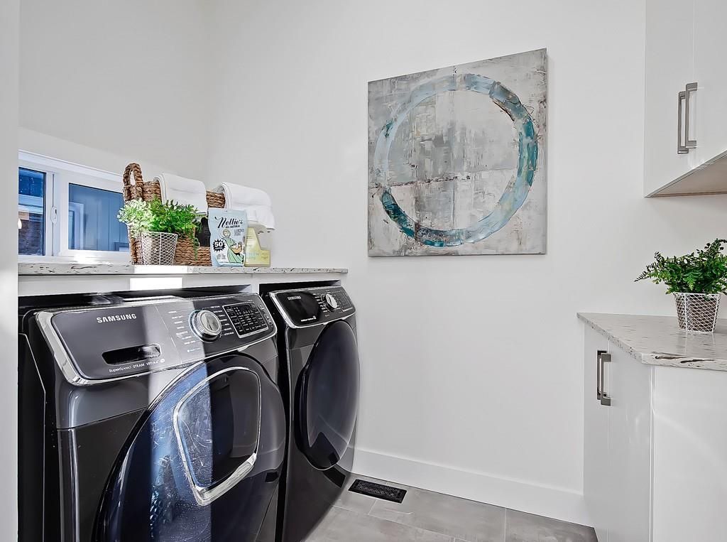 A laundry room with two dark metallic appliances, a white countertop, wall cabinets, and a circular abstract wall art.