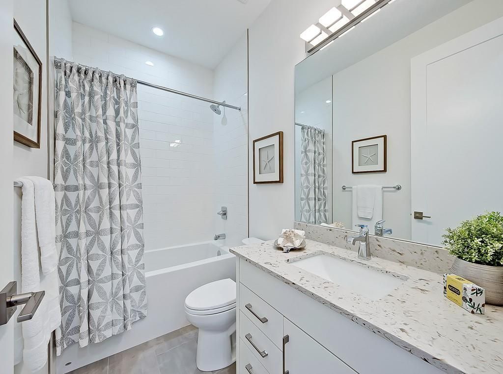 A bright, modern bathroom featuring a white vanity with a speckled countertop, a toilet, and a bathtub with a curtain.