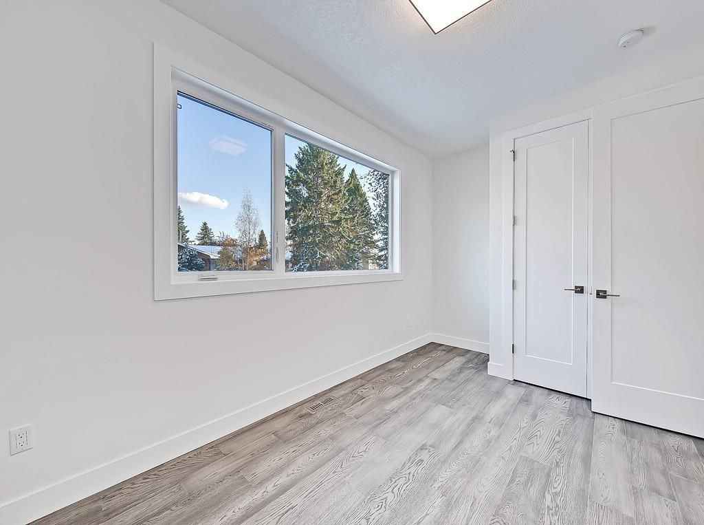 A minimalist room with white walls, light gray wood-grain floors, a large window, and two closed white doors.