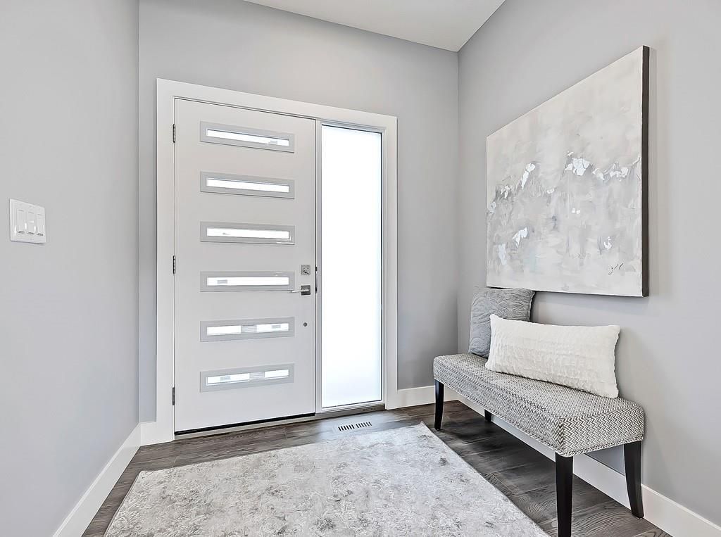 A light-filled foyer with a modern white door with glass panels, a patterned grey bench, and abstract wall art.