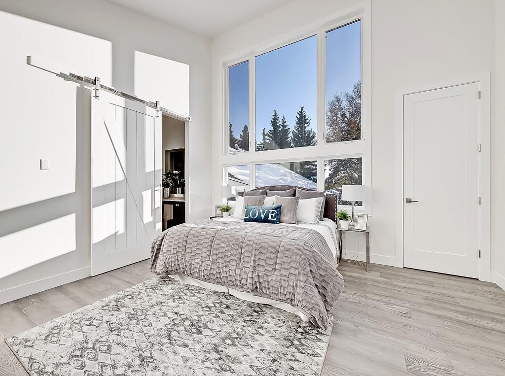 A bright, modern bedroom featuring a bed with a textured grey blanket, a large window, and a white sliding barn door.