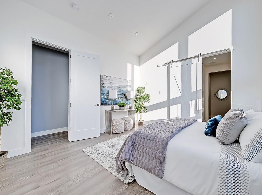 A bright, modern bedroom featuring a neatly made bed with grey bedding, light wood floors, and floor-to-ceiling windows.