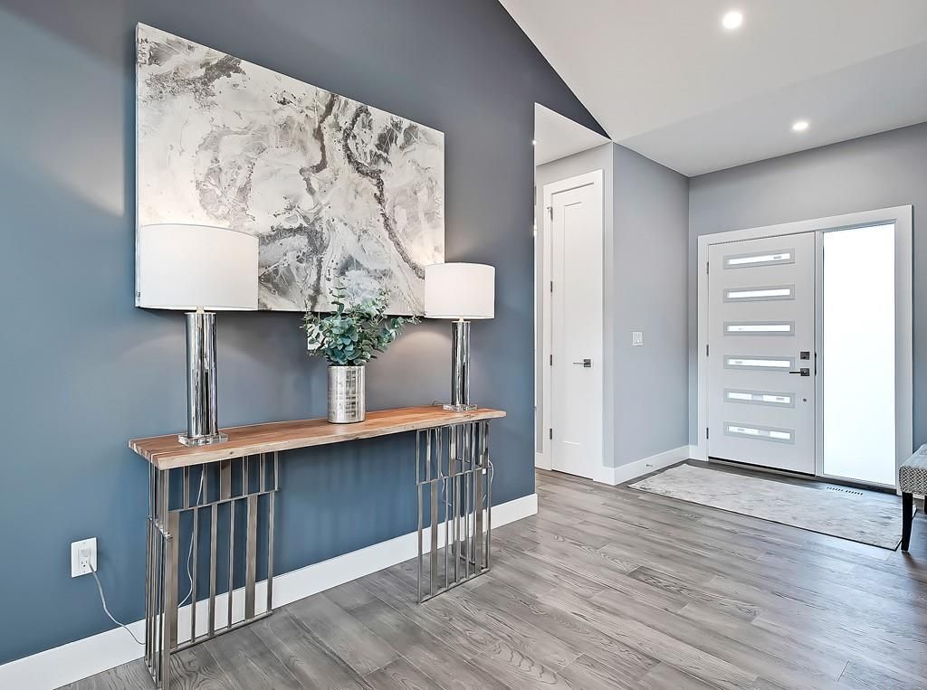 A modern foyer features a metal console table with two lamps and an abstract painting against a blue wall near a white door.