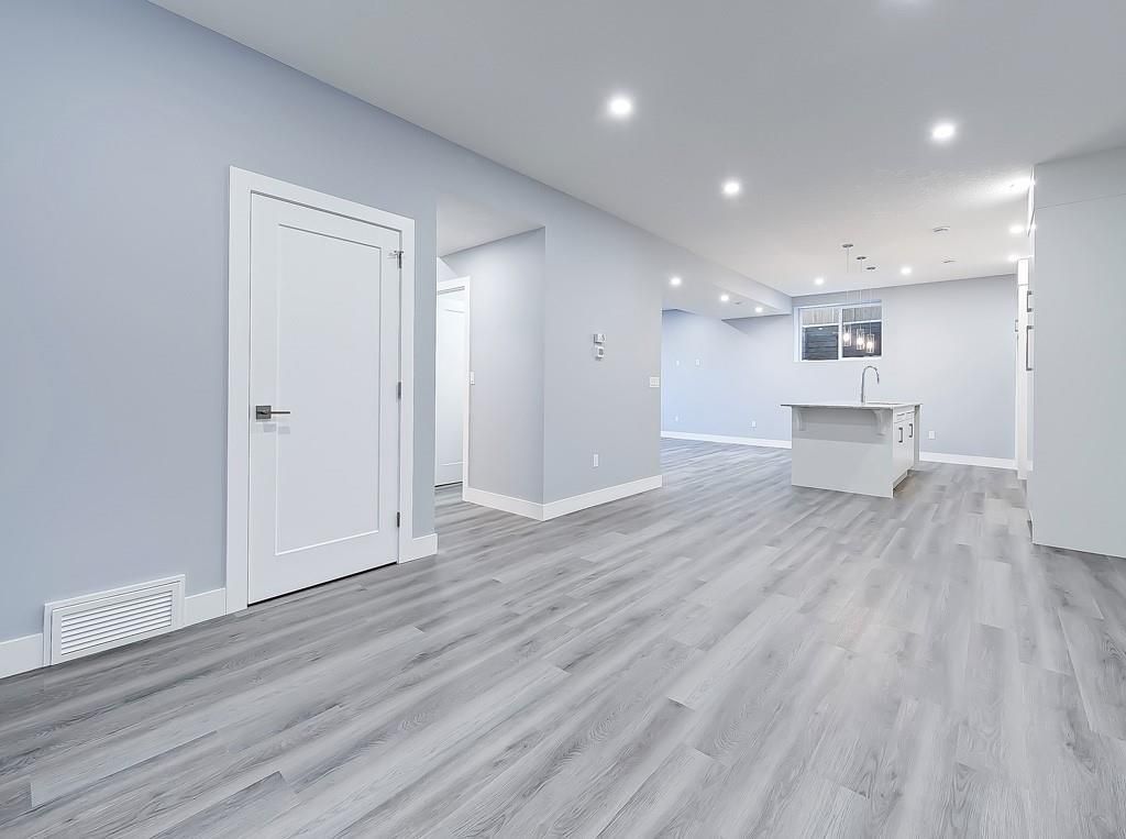 A bright, unfurnished basement room with light grey walls, grey wood-look flooring, a white door, and a kitchen island.