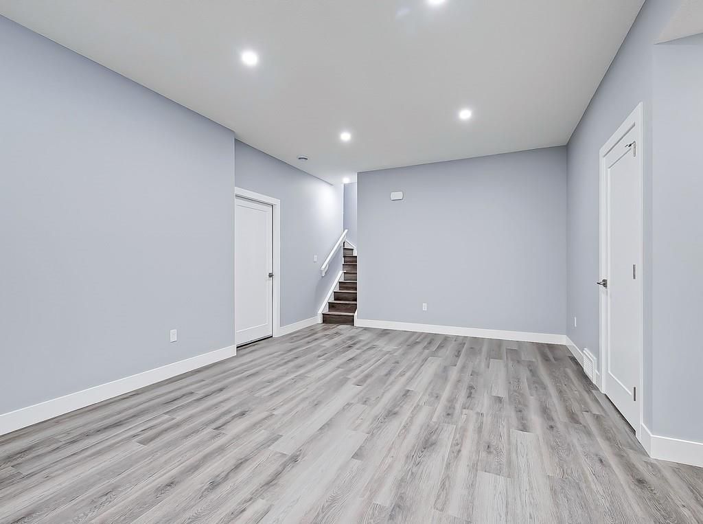 A bright, empty room with light gray walls, white trim, and gray wood-look flooring, featuring stairs in the background.