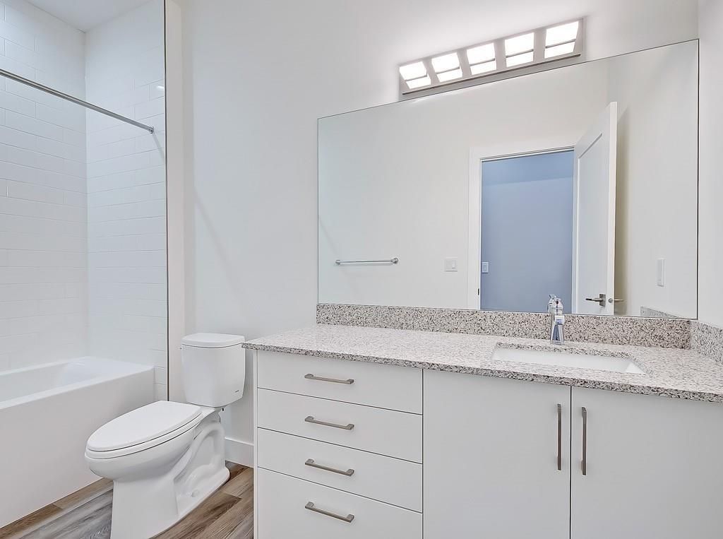 A modern bathroom with a white vanity, a large mirror, a toilet, and a white bathtub with a tiled surround.