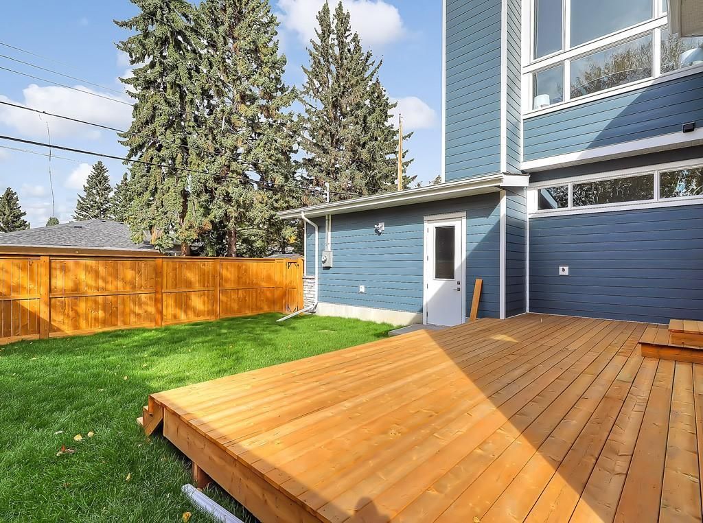 A wooden deck adjacent to a blue house with a white door and a backyard lawn enclosed by a tall wooden fence.