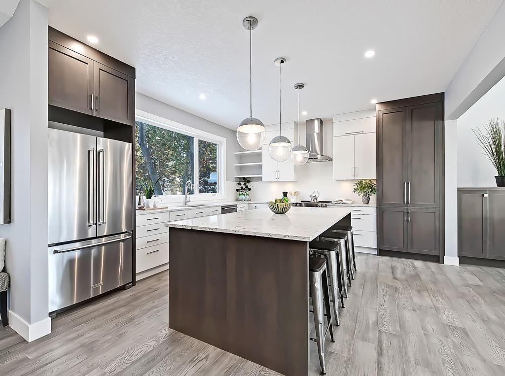 A modern kitchen featuring a dark wooden island with bar stools, white countertops, stainless appliances, and pendant lights.