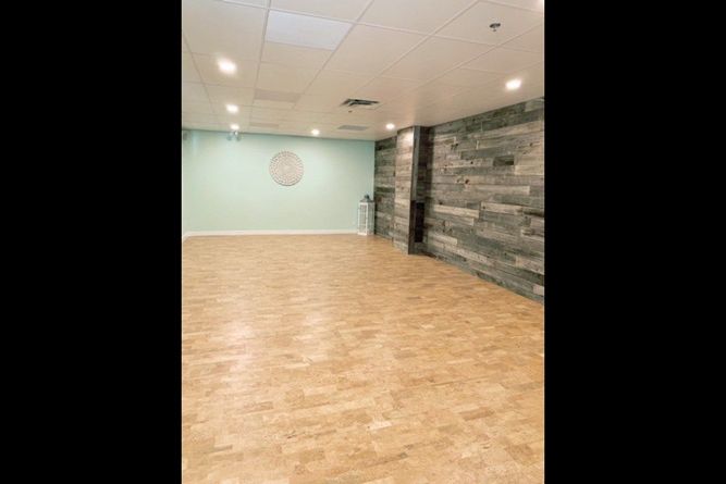 An empty room with light wood floors, a soft green end wall, and a textured grey wood-paneled side wall under recessed lights.