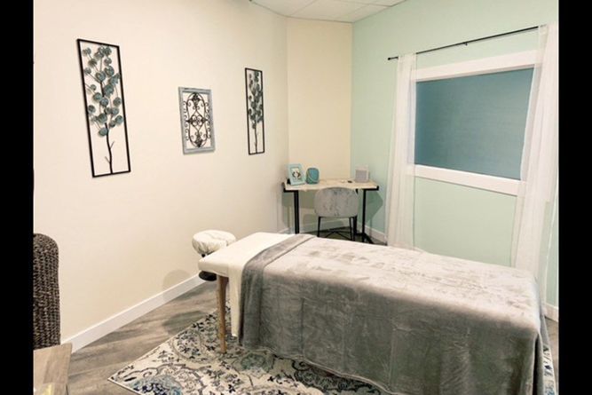 A minimalist treatment room with a massage table, grey rug, desk, chair, and decorative wall art on cream and teal walls.