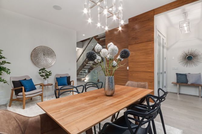A modern dining room with a wooden table, black chairs, a glass vase of flowers, and a staircase in the background.
