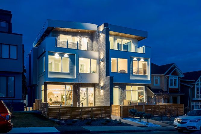 A modern multi-story residential building at twilight, featuring illuminated glass windows and a wooden fence.