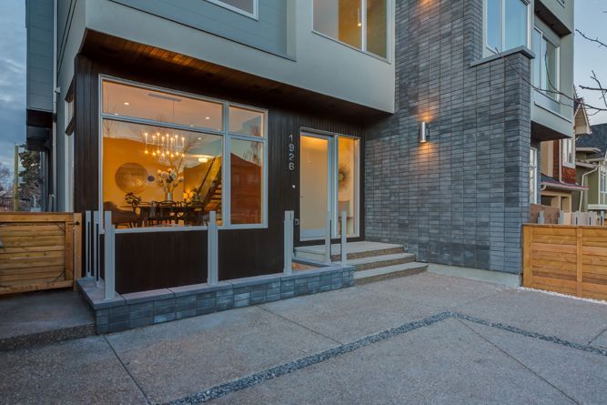 Modern house exterior featuring a stone-clad wall, large window with interior lighting, and a paved front entrance.