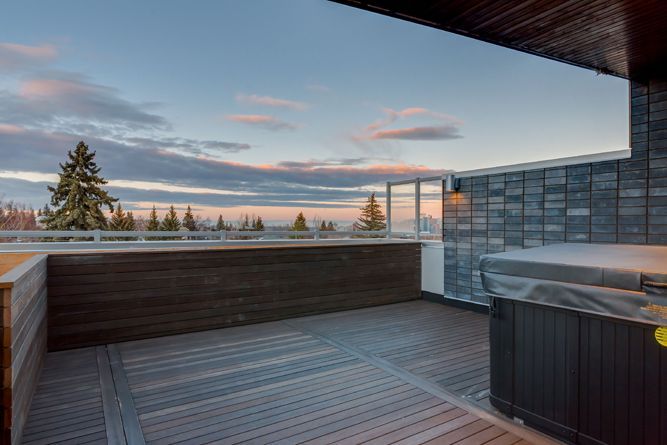 A modern outdoor wooden deck at sunset featuring a covered hot tub, glass railing, and a scenic view of evergreen trees.