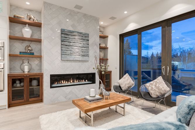 A modern living room with a linear fireplace, marble wall, wood shelves, coffee table, and two chairs near glass doors.