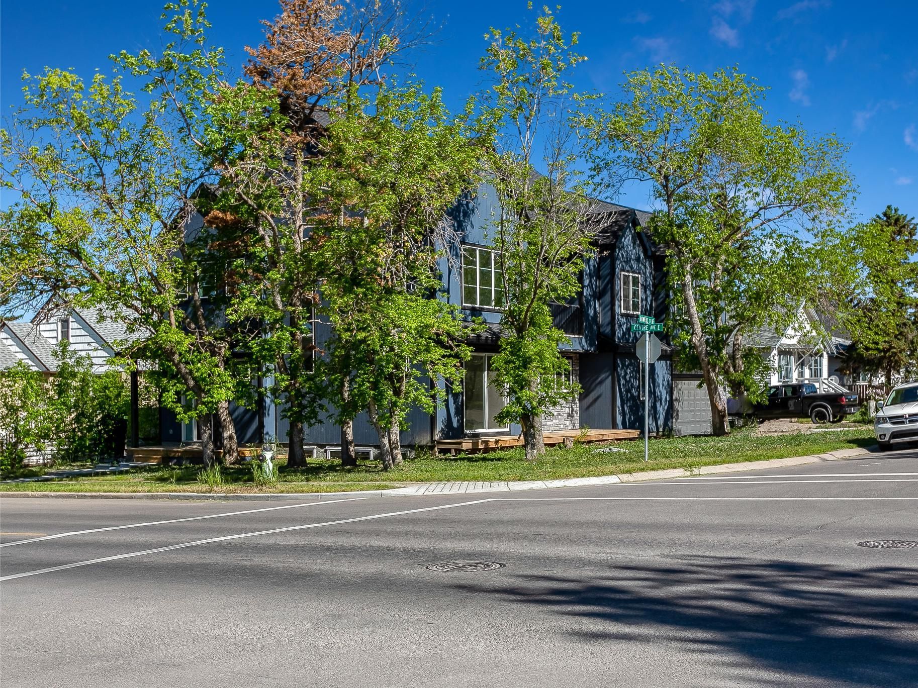 A two-story blue house sits behind lush green trees next to a paved road on a bright, sunny day.