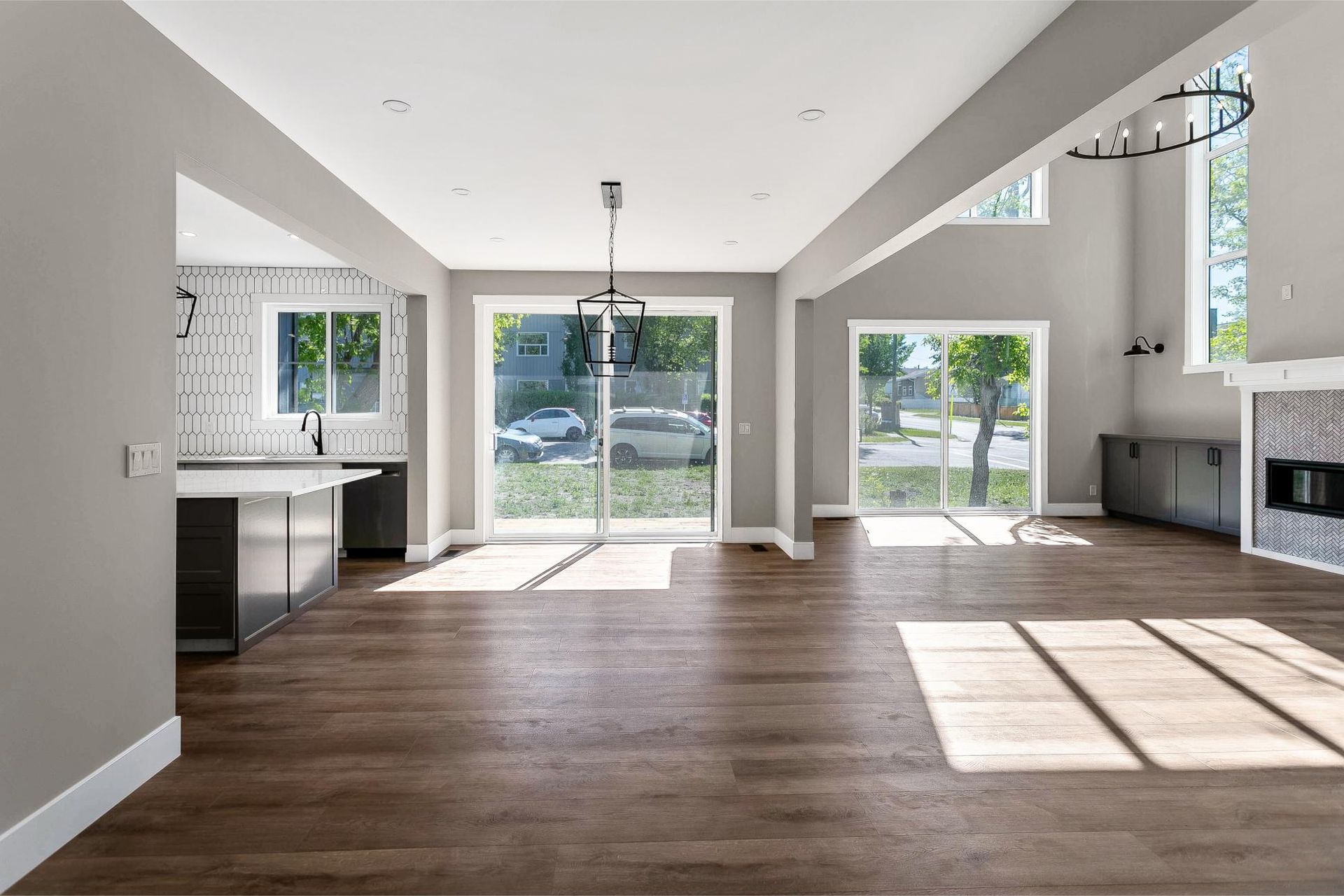 A sunlit, open-concept living area with grey walls, dark wood floors, a kitchen island, and a stone fireplace.