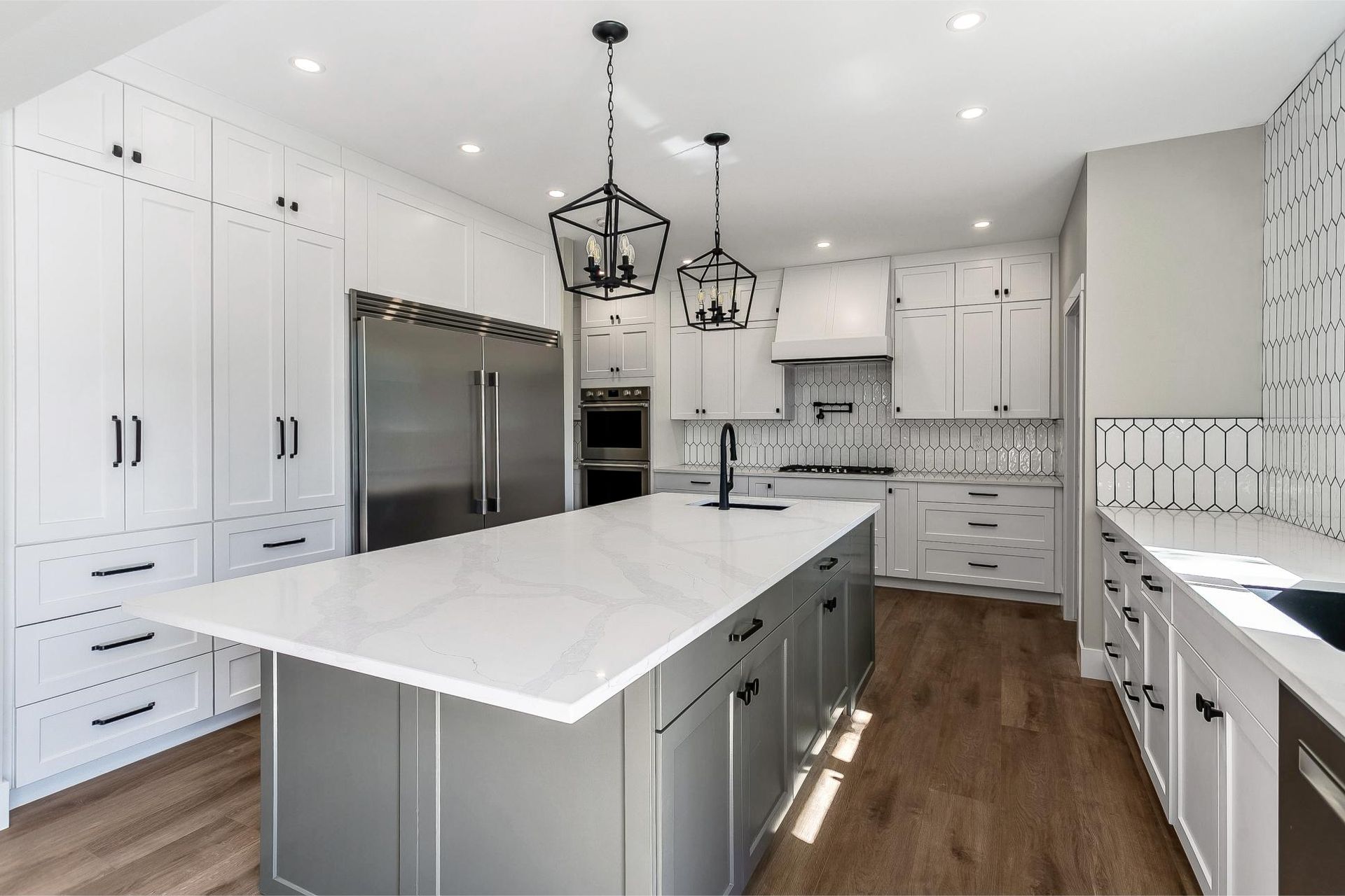 A modern kitchen with a large gray island, white cabinets, stainless steel refrigerator, and wood-look flooring.