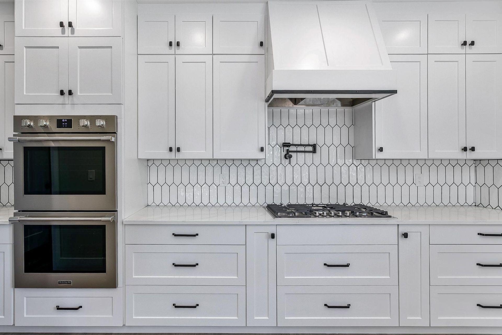 A modern white kitchen featuring a double wall oven, a gas cooktop, patterned tile backsplash, and dark hardware.
