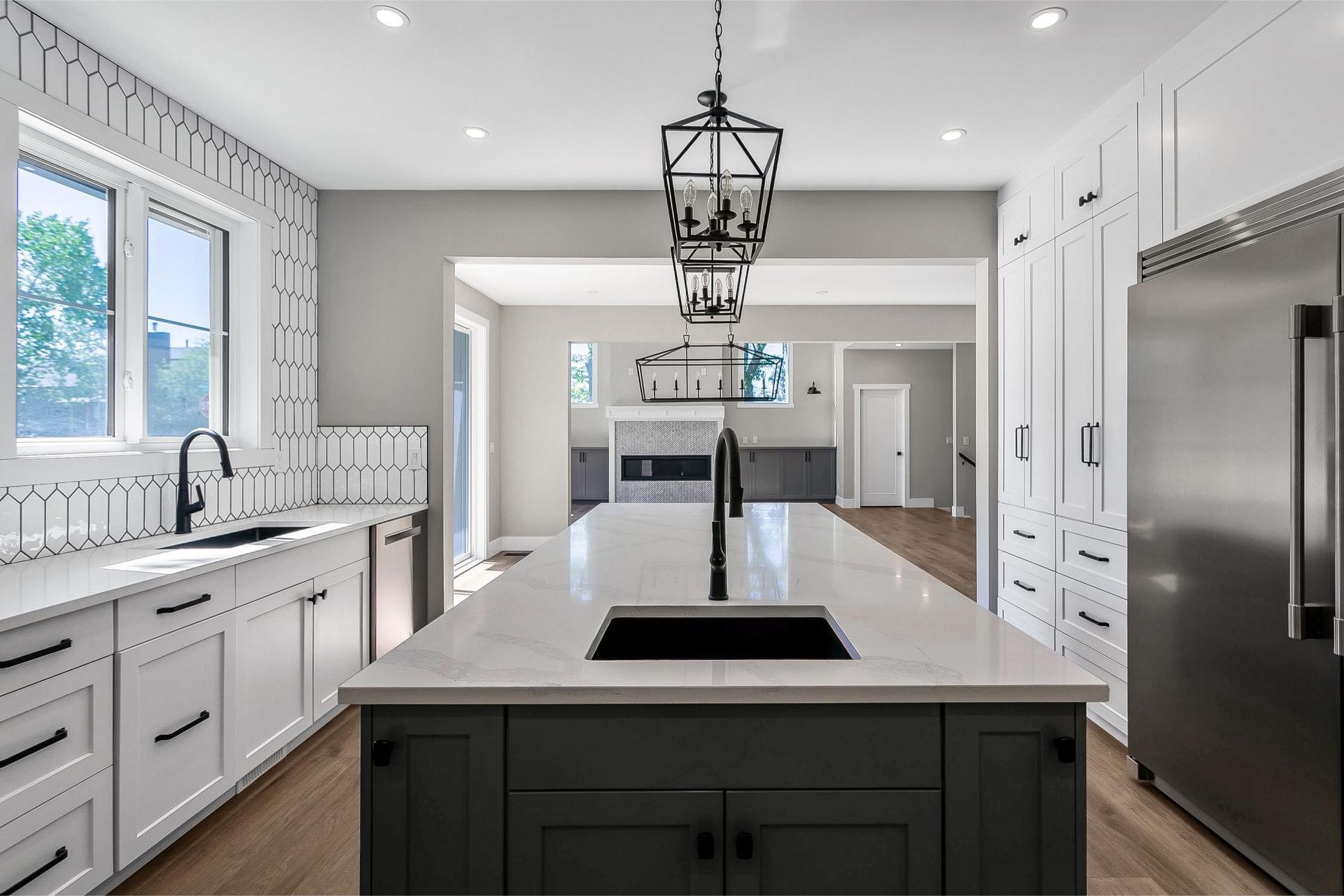A bright, modern kitchen featuring white cabinets, a dark grey central island with a sink, and a black geometric pendant.