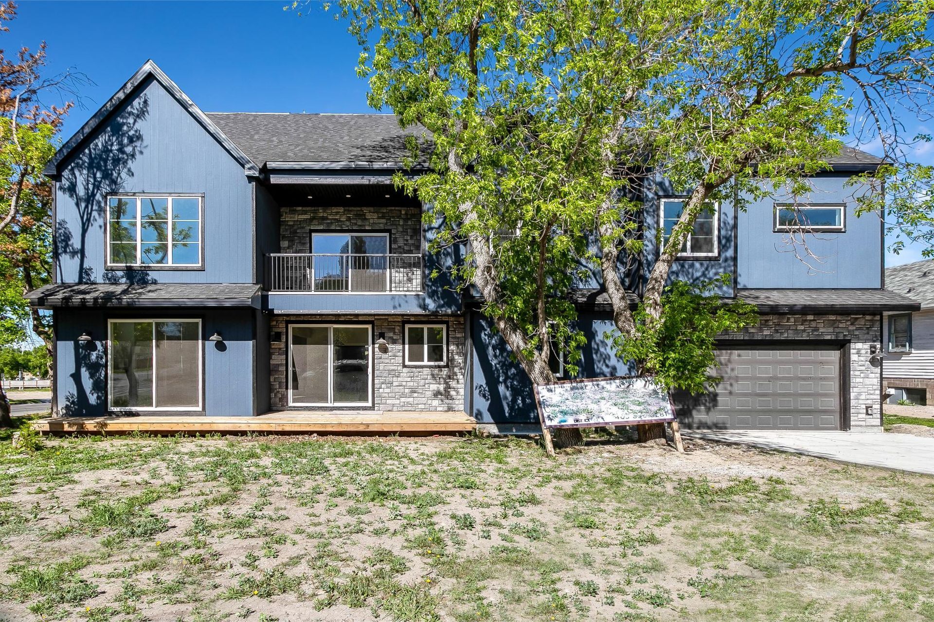 A two-story blue house with stone accents, a garage, and a large tree in the front yard under a clear blue sky.