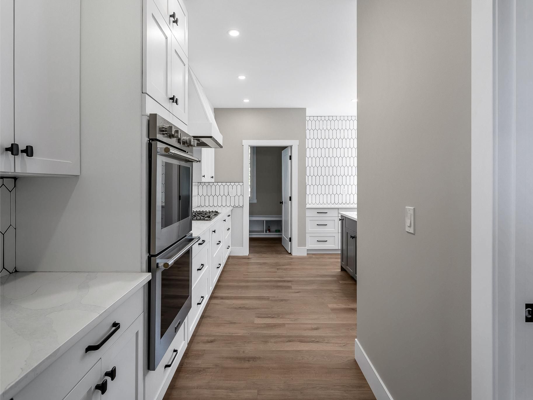 A modern kitchen featuring white cabinets, stainless steel double ovens, wooden floors, and a doorway to another room.