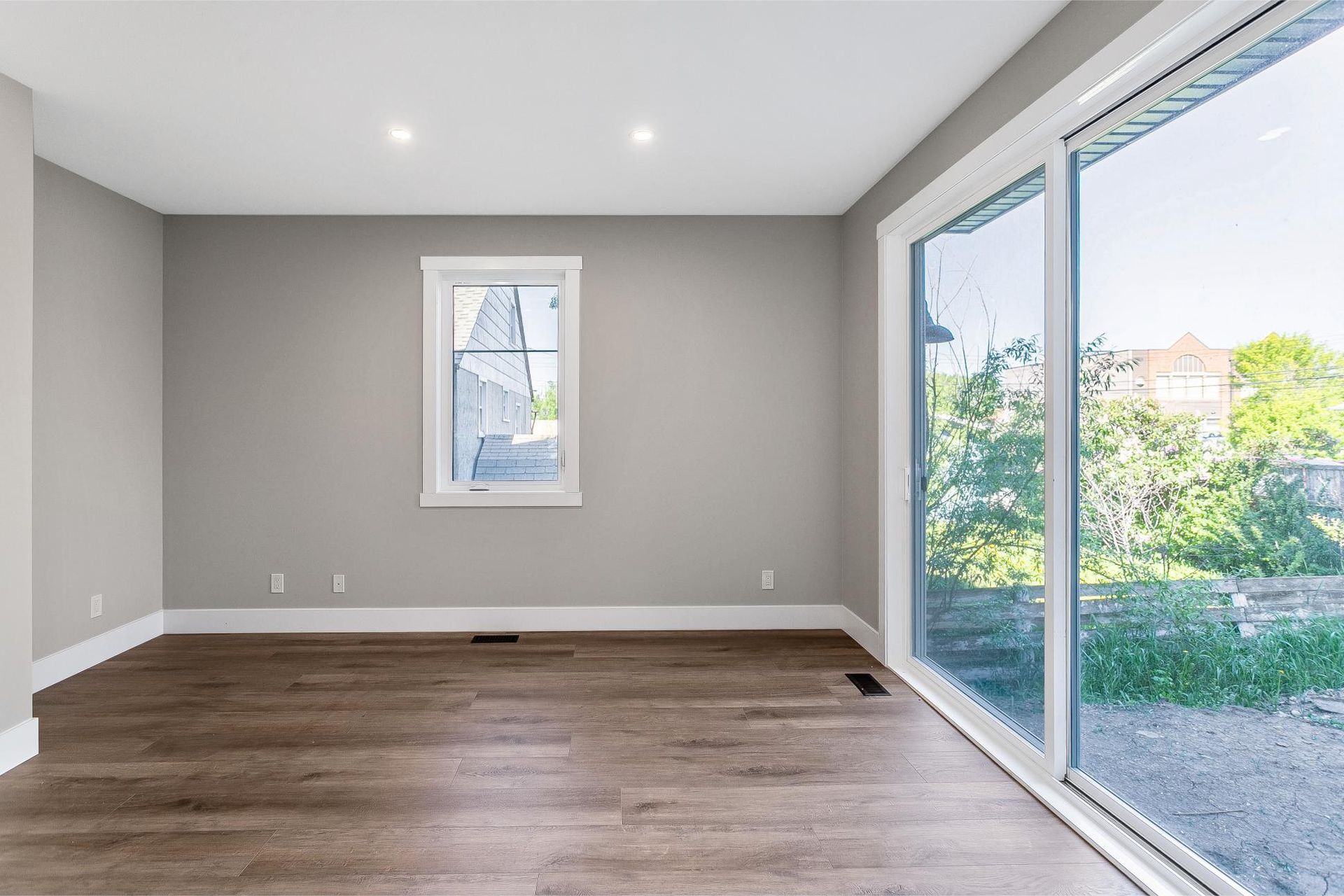 An empty room with light gray walls, dark wood floors, a small window, and a large sliding glass door.
