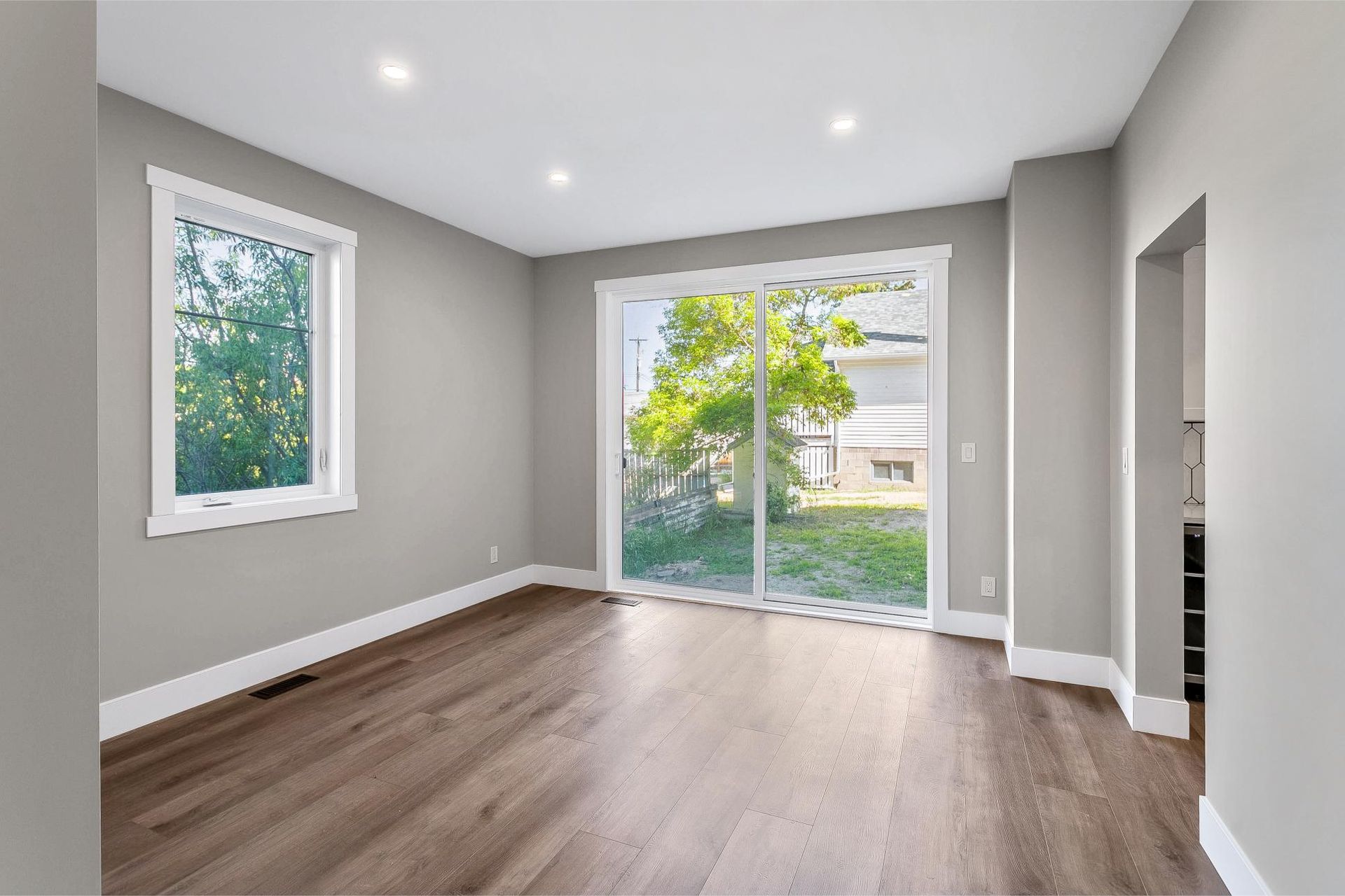 An empty room with light gray walls, dark wood-look flooring, a window, and sliding glass doors leading to a backyard.