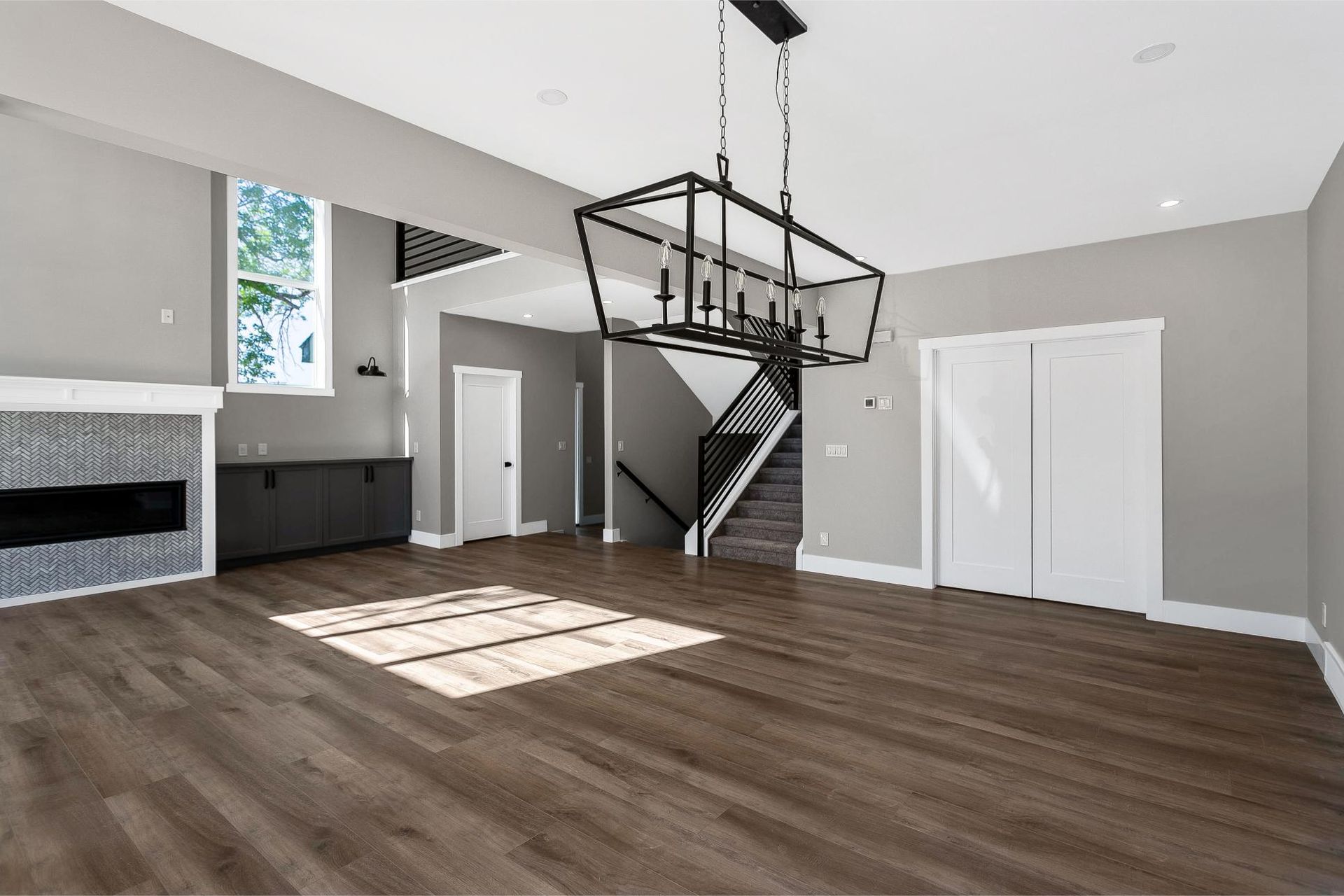 Modern living room with wood floors, gray walls, a linear fireplace, staircase, and a large black metal chandelier.