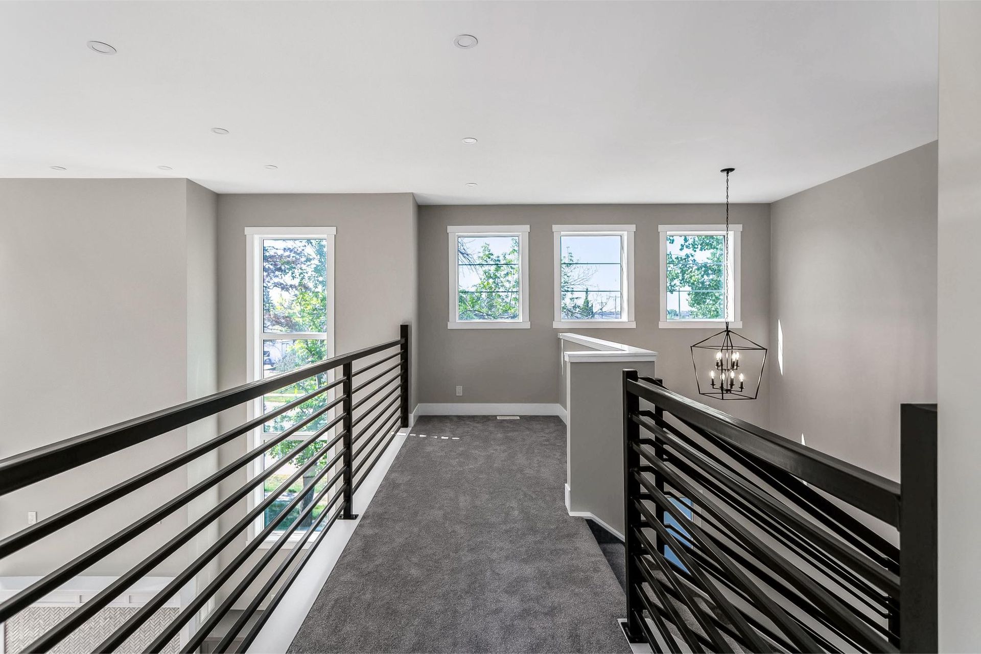 An indoor balcony hallway with dark railings, grey carpet, and three large windows along a light grey wall.