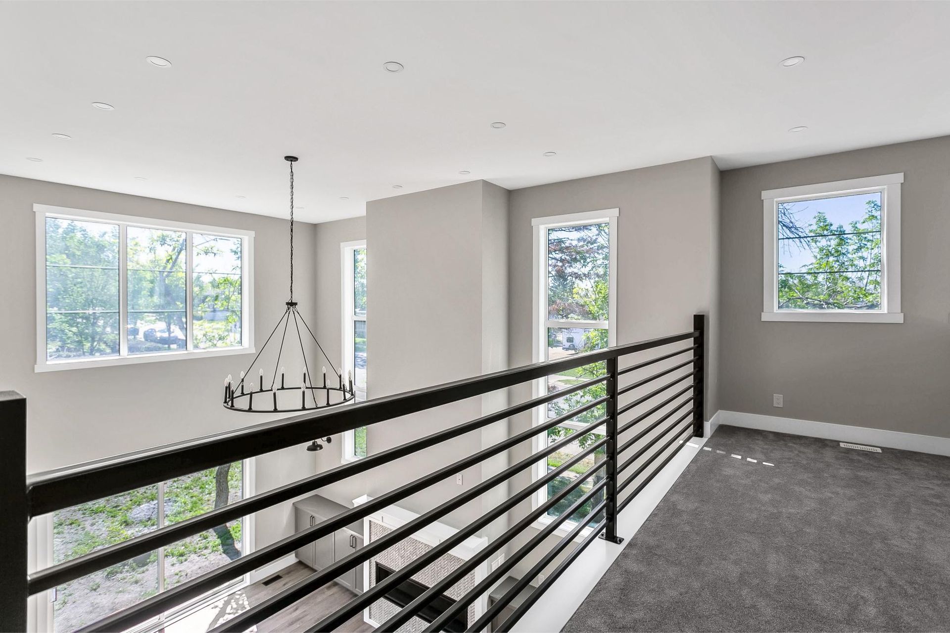 An elevated view from a carpeted hallway looking down over a black railing into a bright, modern, open-concept living area.