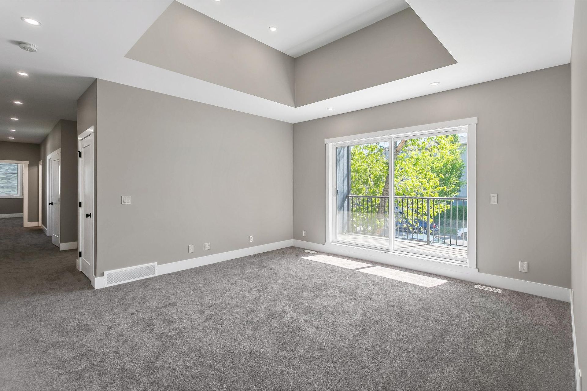 Empty, carpeted room with light gray walls, a tray ceiling with recessed lighting, and a large sliding glass door.